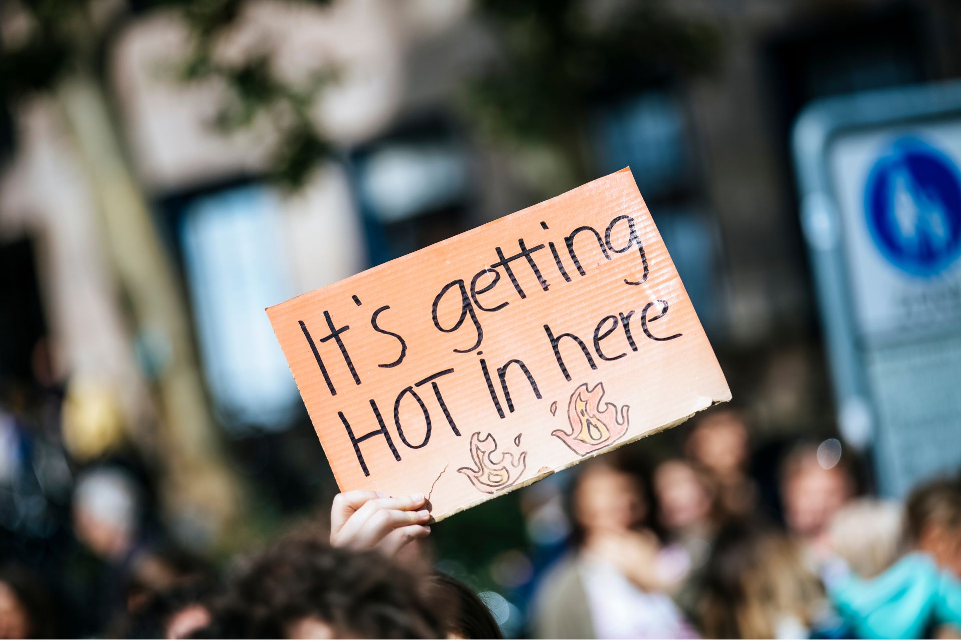 Person in a crowd holding up a handwritten cardboard protest sign reading "It's getting HOT in here."