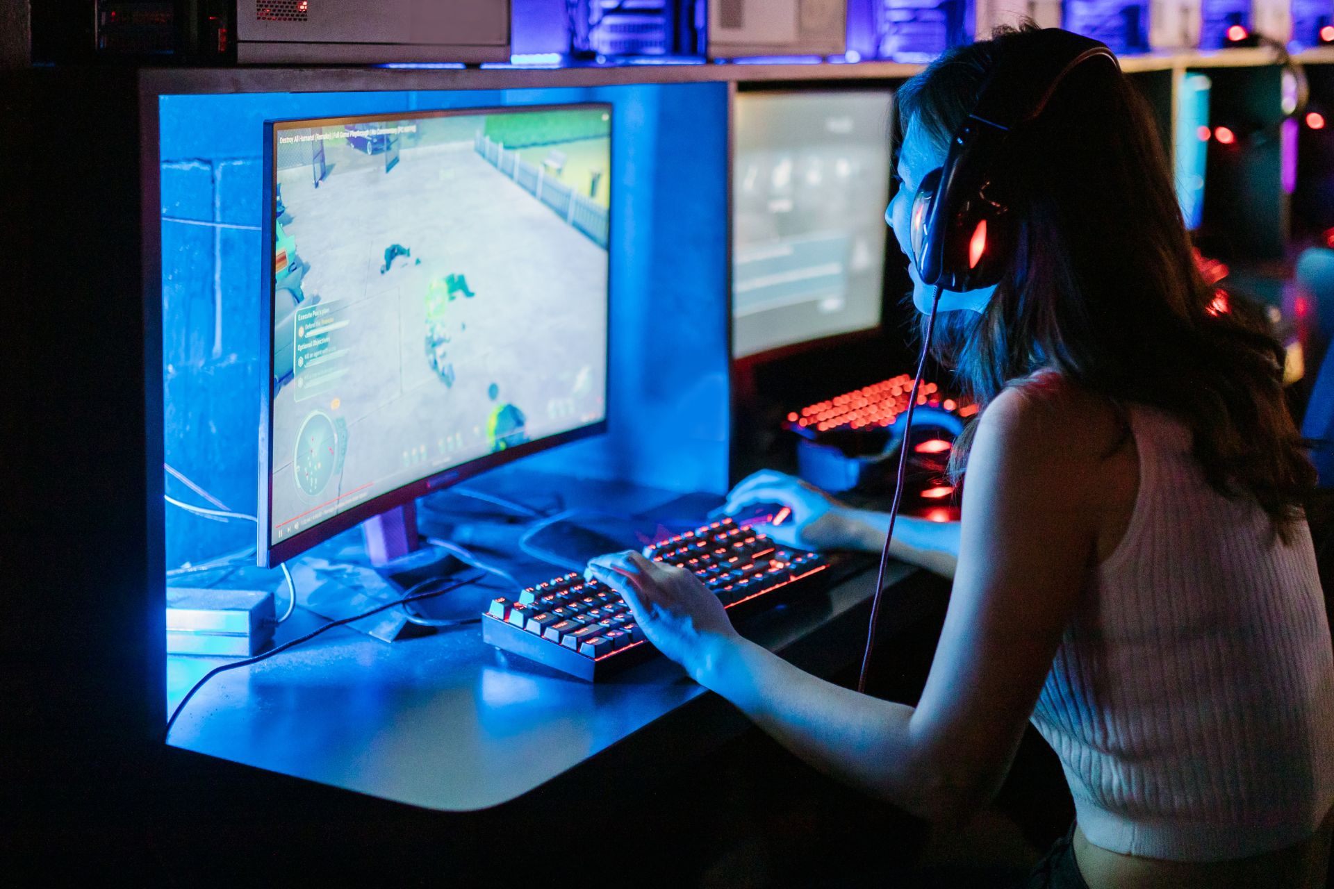 Young woman wearing a headset, playing a video game in front of dual monitors in a room lit by blue neon light.