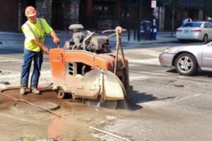 Construction worker in a high-visibility safety vest operating a walk-behind concrete saw on a city street.