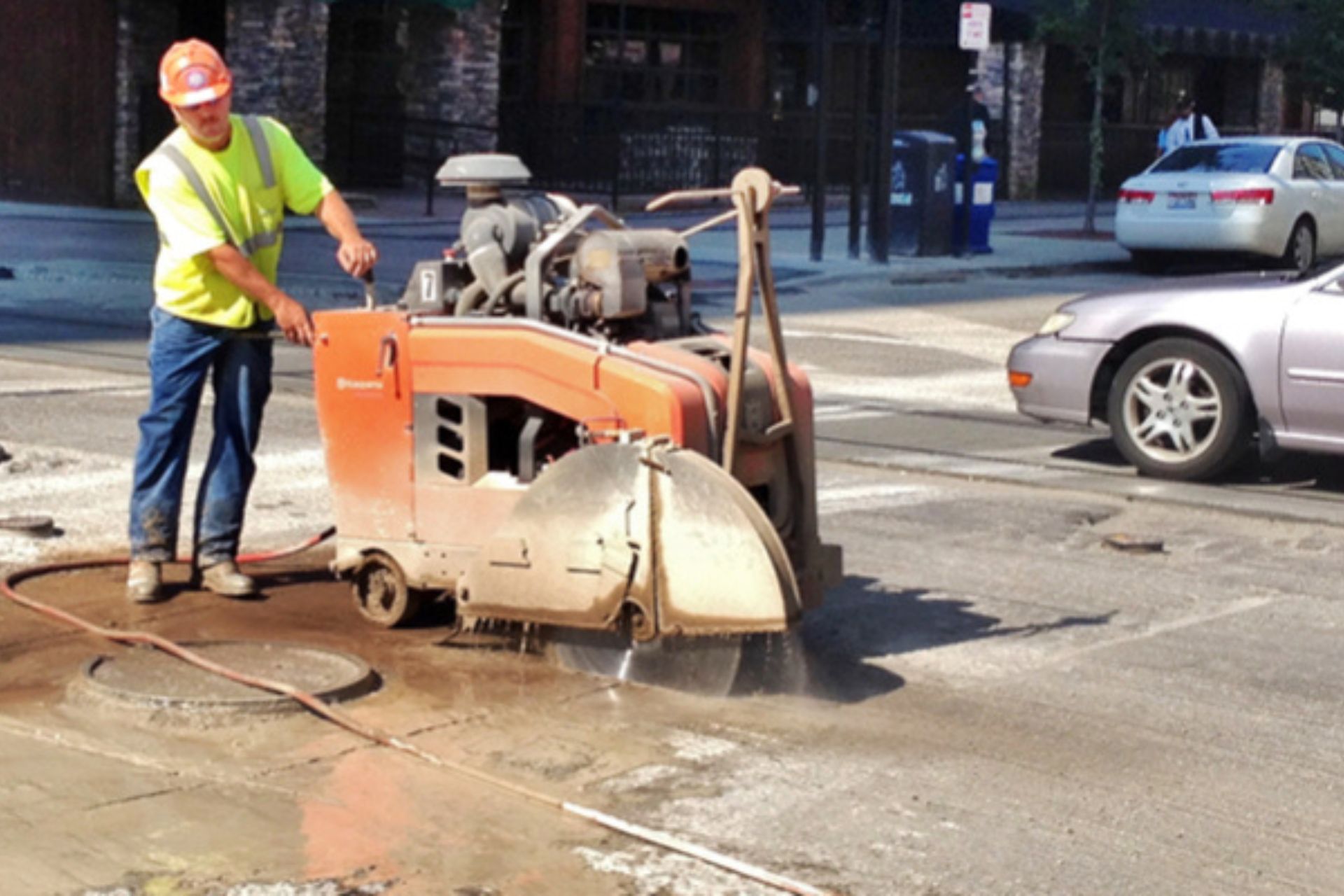 Construction worker in a high-visibility safety vest operating a walk-behind concrete saw on a city street.