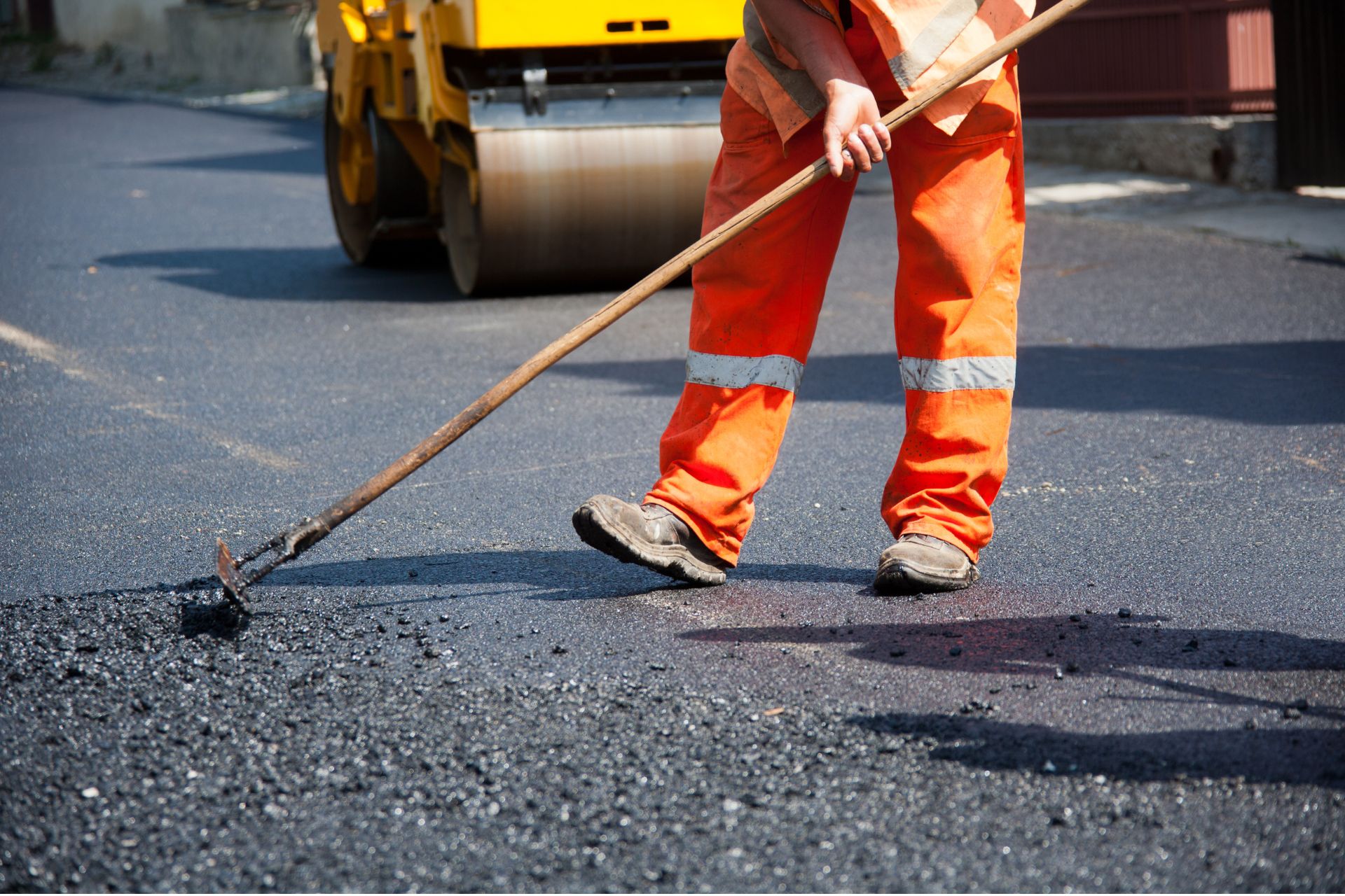 A construction worker in high-visibility orange pants spreading fresh asphalt on a road.
