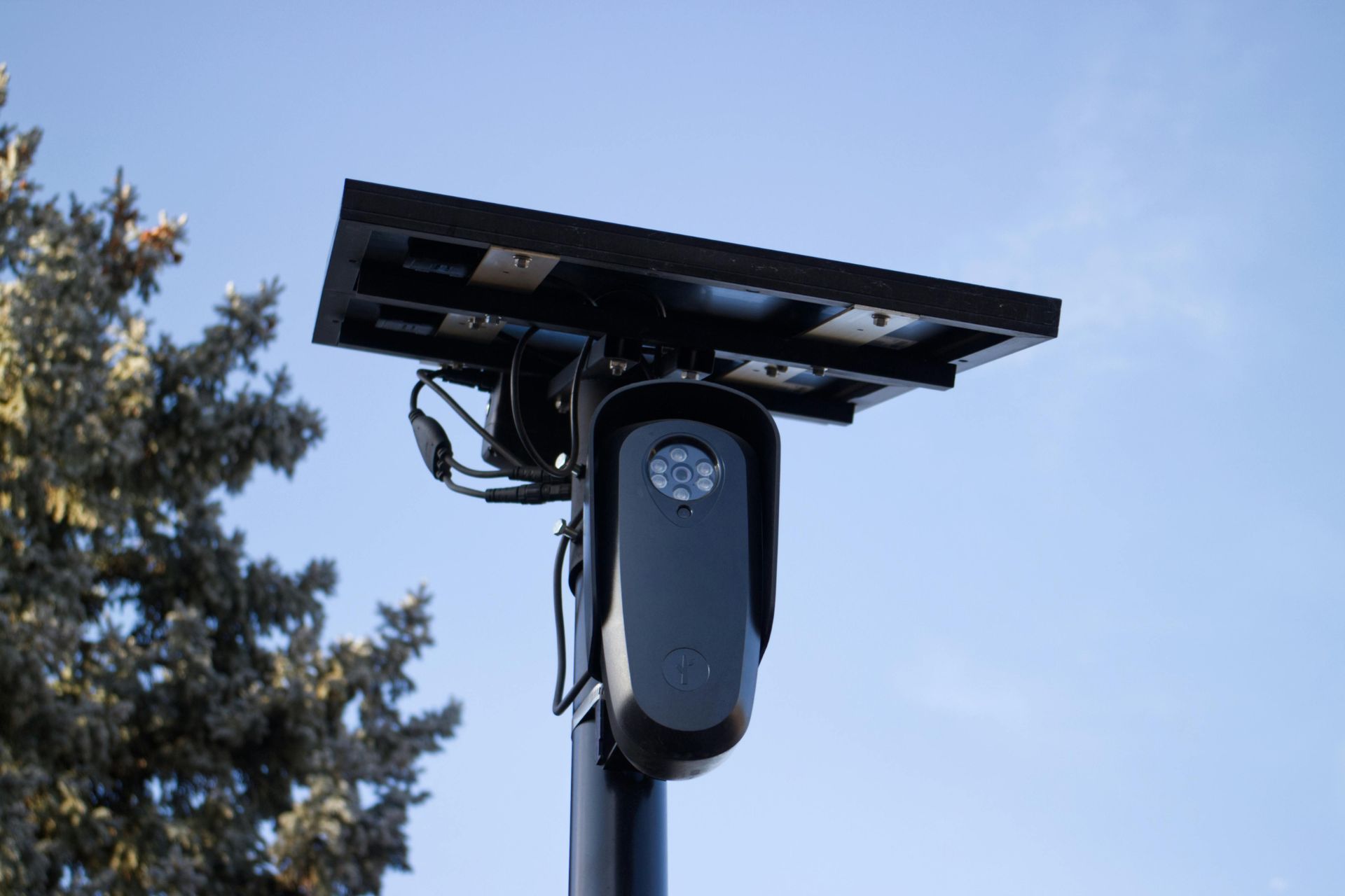 A solar-powered surveillance camera mounted on a black pole against a clear blue sky.