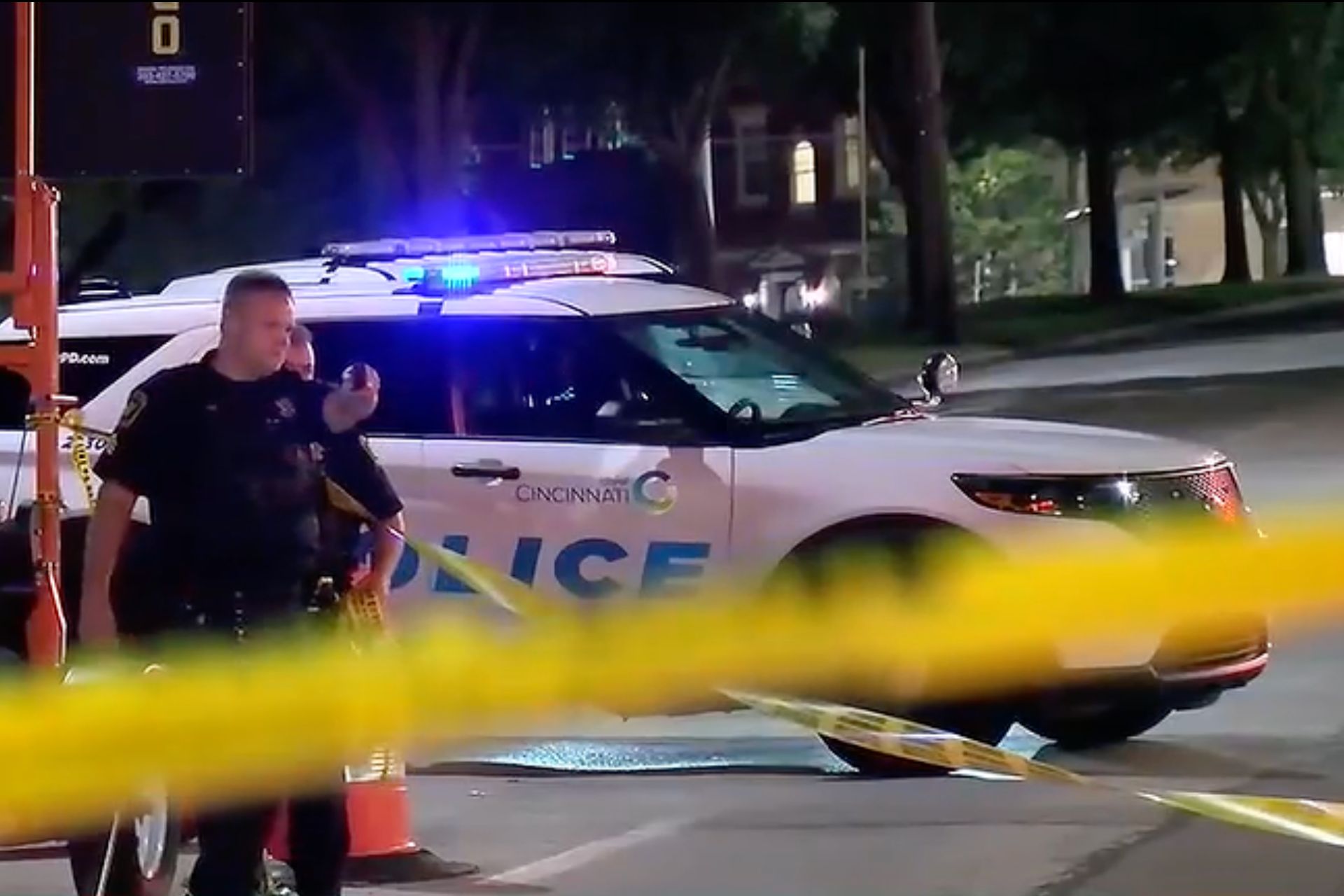 A police officer standing near a patrol car with flashing emergency lights behind yellow caution tape.