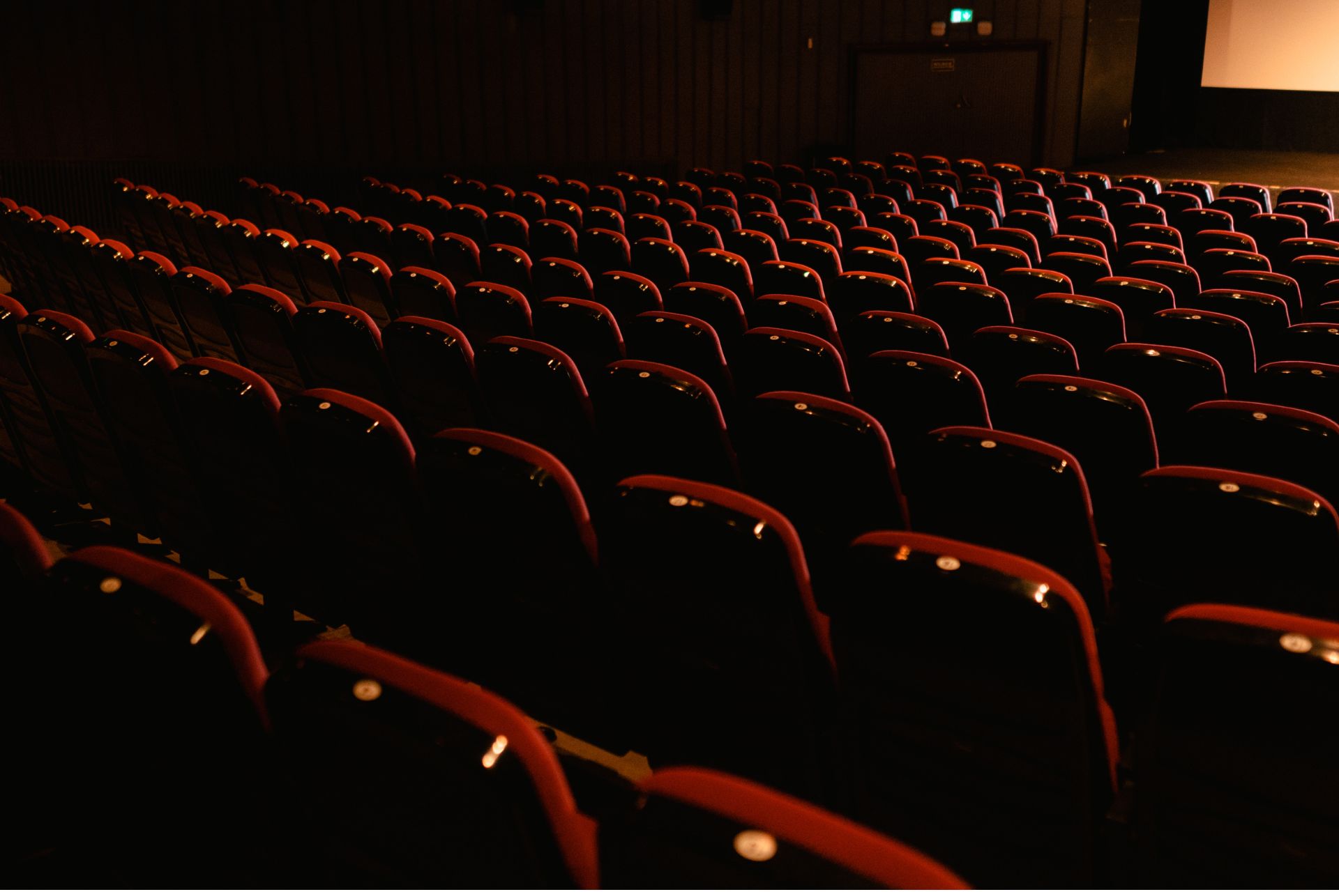 Rows of empty red plush seats inside a dark movie theater auditorium facing a large screen.