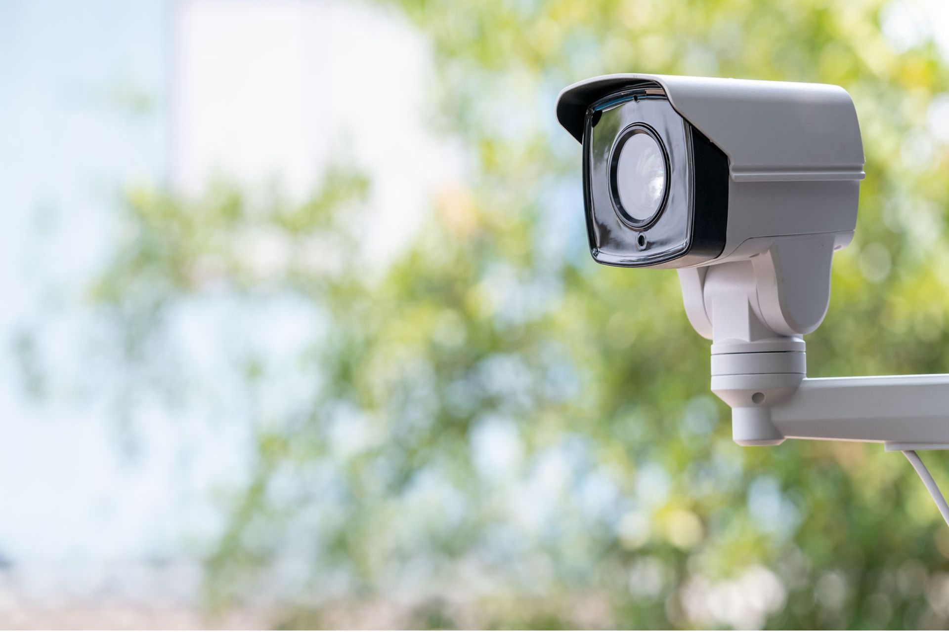 Outdoor security surveillance camera mounted with green leafy trees in the background.