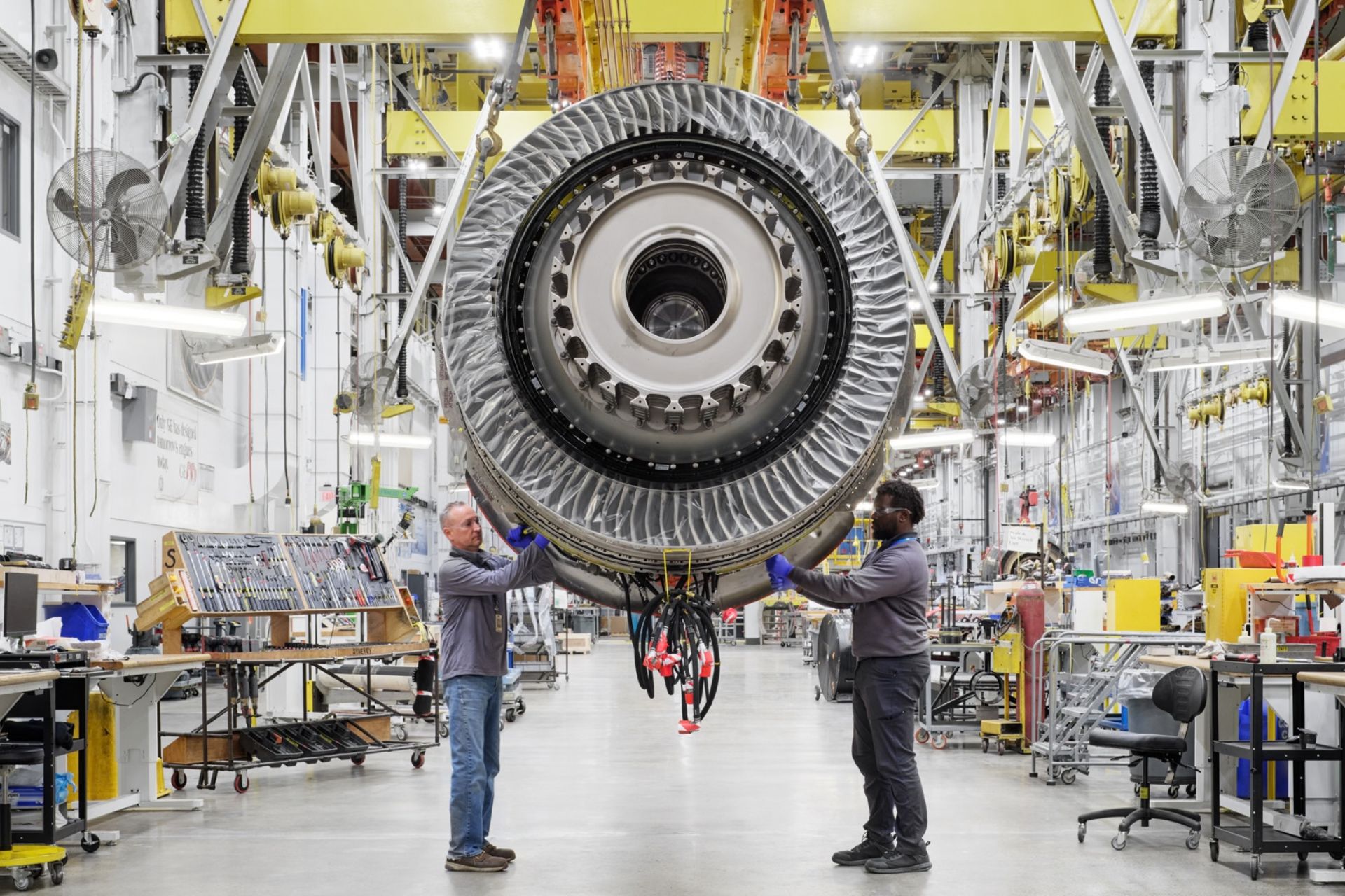 Two technicians working on a large jet engine suspended inside a GE Aerospace manufacturing facility.