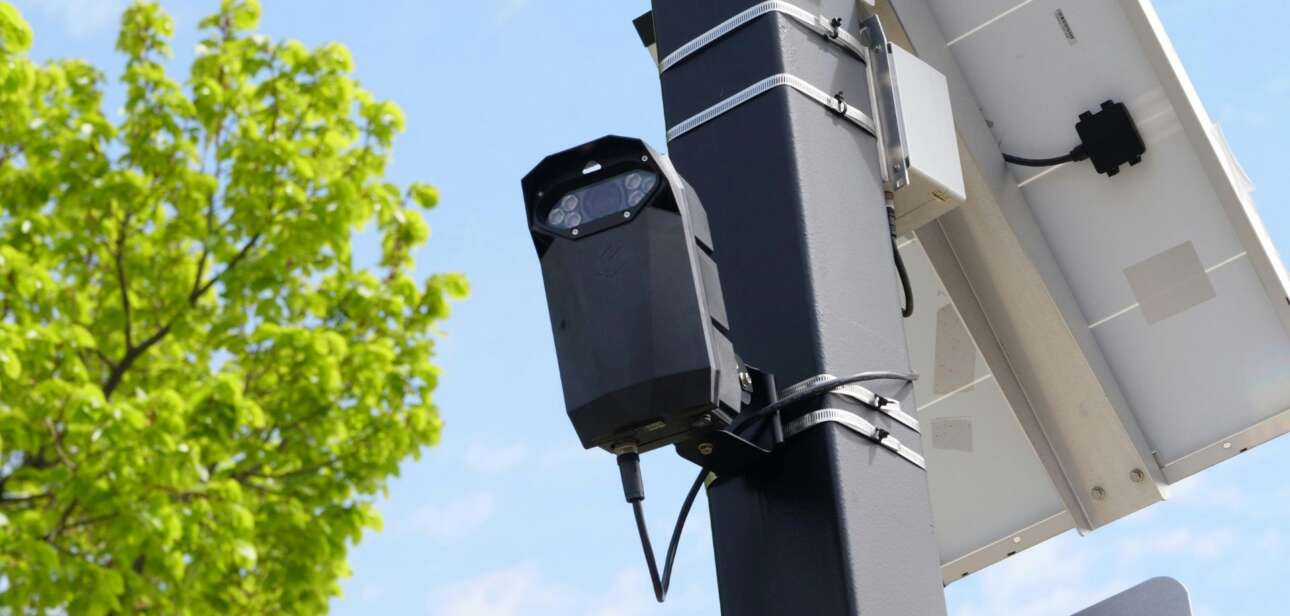 A black Flock safety camera and solar panel mounted on a utility pole against a blue sky.