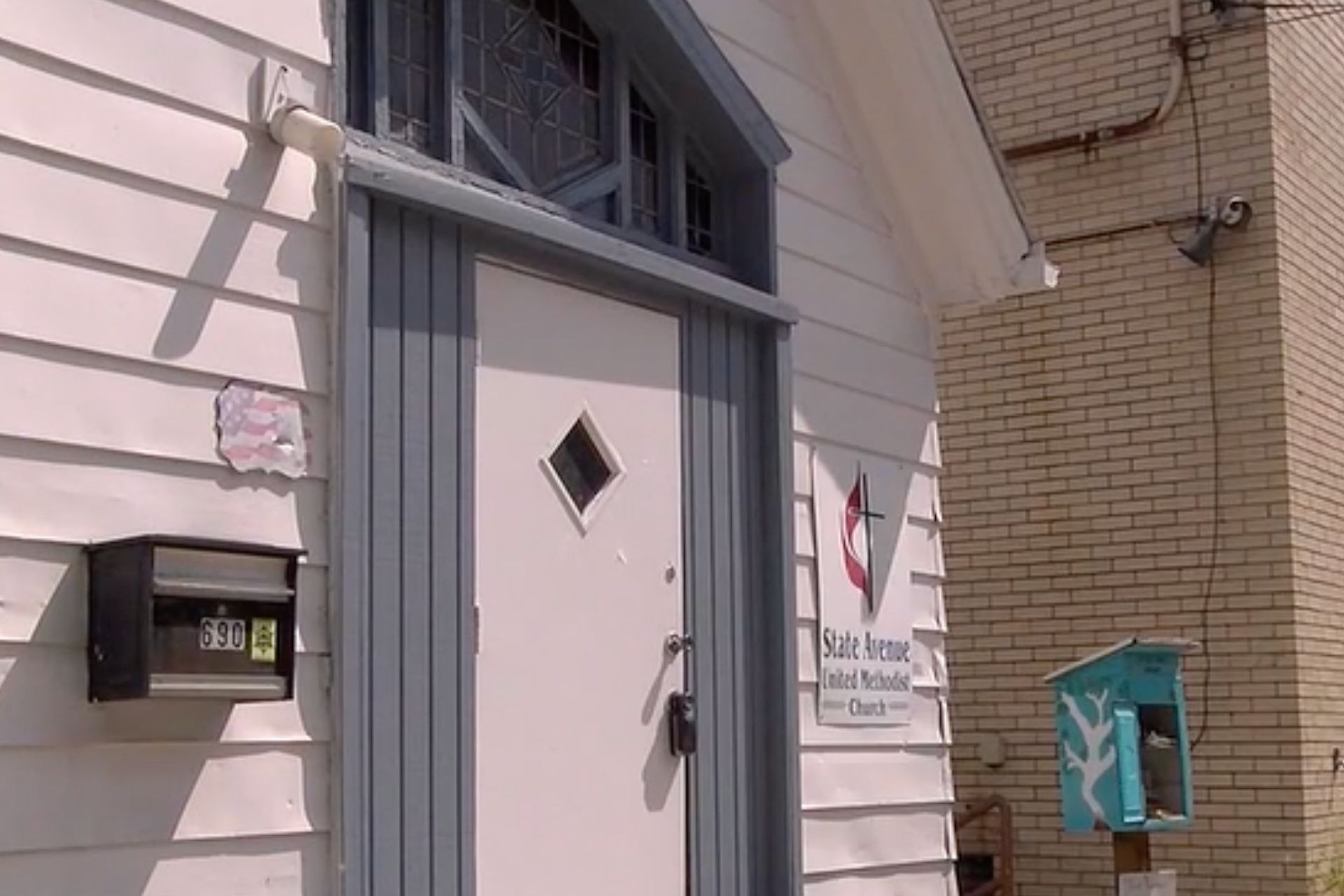 The exterior entrance of a church featuring white siding, a grey door with a diamond window, and a wall-mounted mailbox.