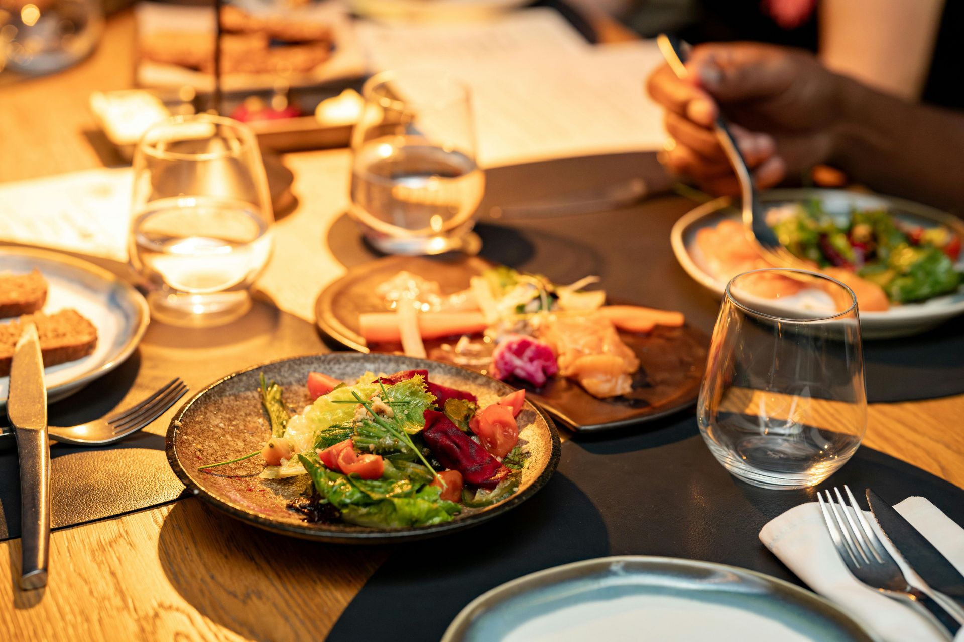 A warmly lit wooden dining table filled with plates of food, wine glasses, and small candles at a restaurant.
