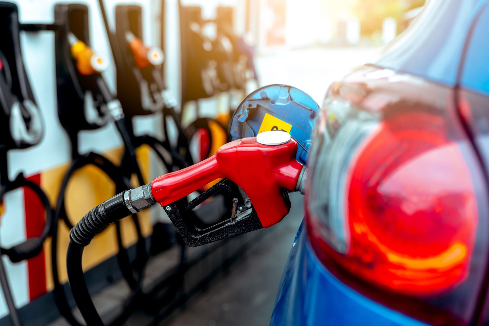 Close-up of the rear of a blue car at a filling station with multiple red pumps in the background.