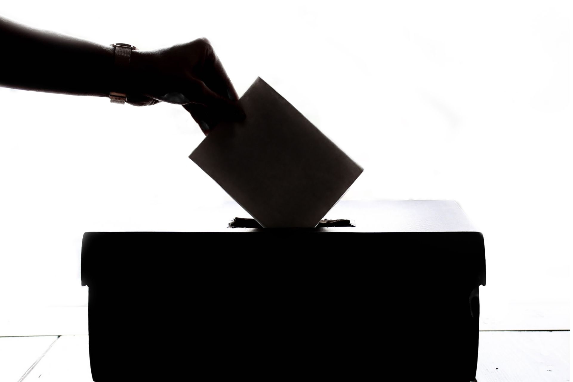 Silhouette of a hand inserting a paper ballot into a voting box slot.