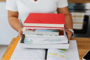 A person holding a tall stack of colorful textbooks and notebooks over a wooden desk.