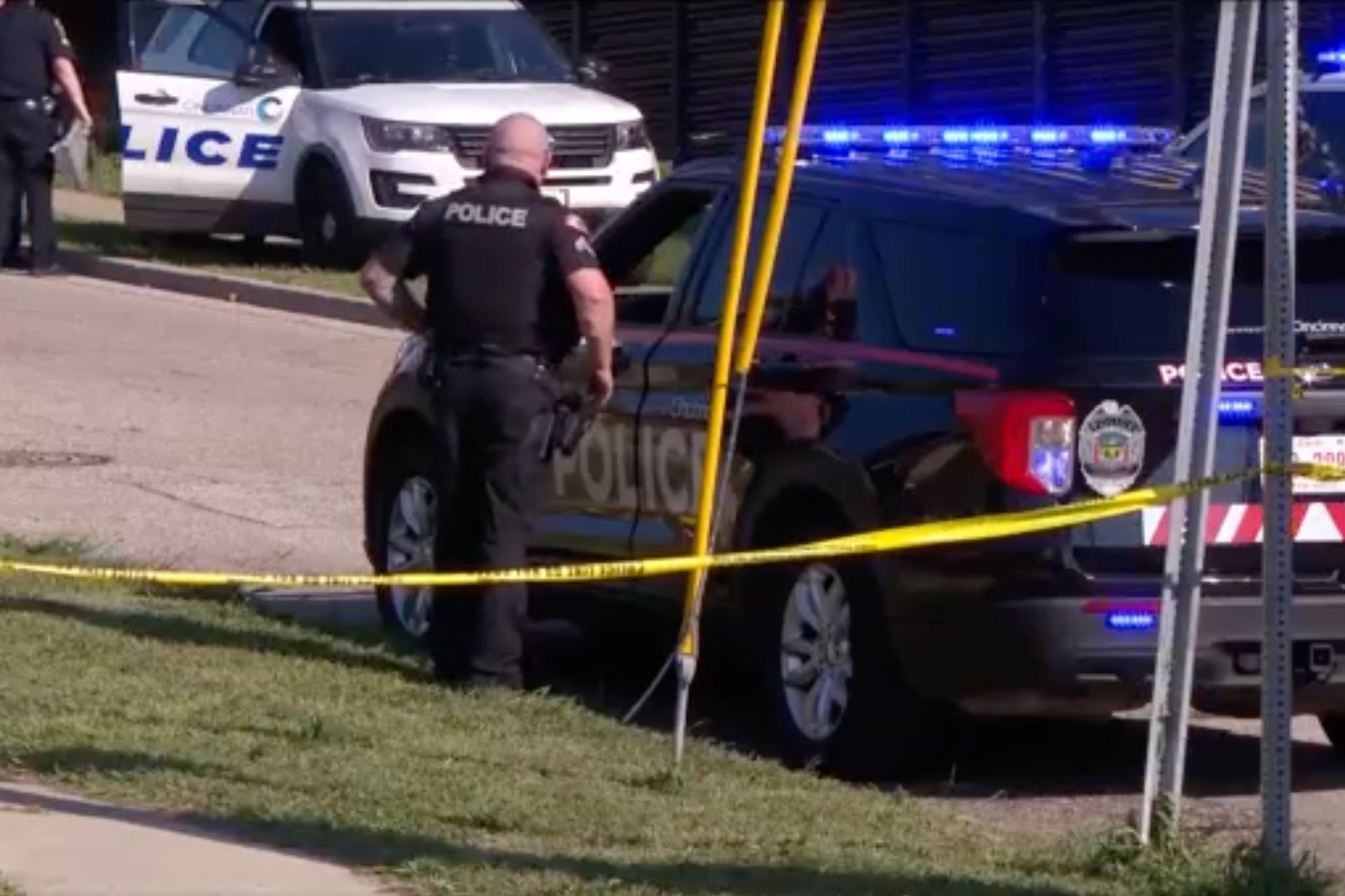 A police vehicle is cordoned off with yellow police tape. An officer stands nearby on the grass.