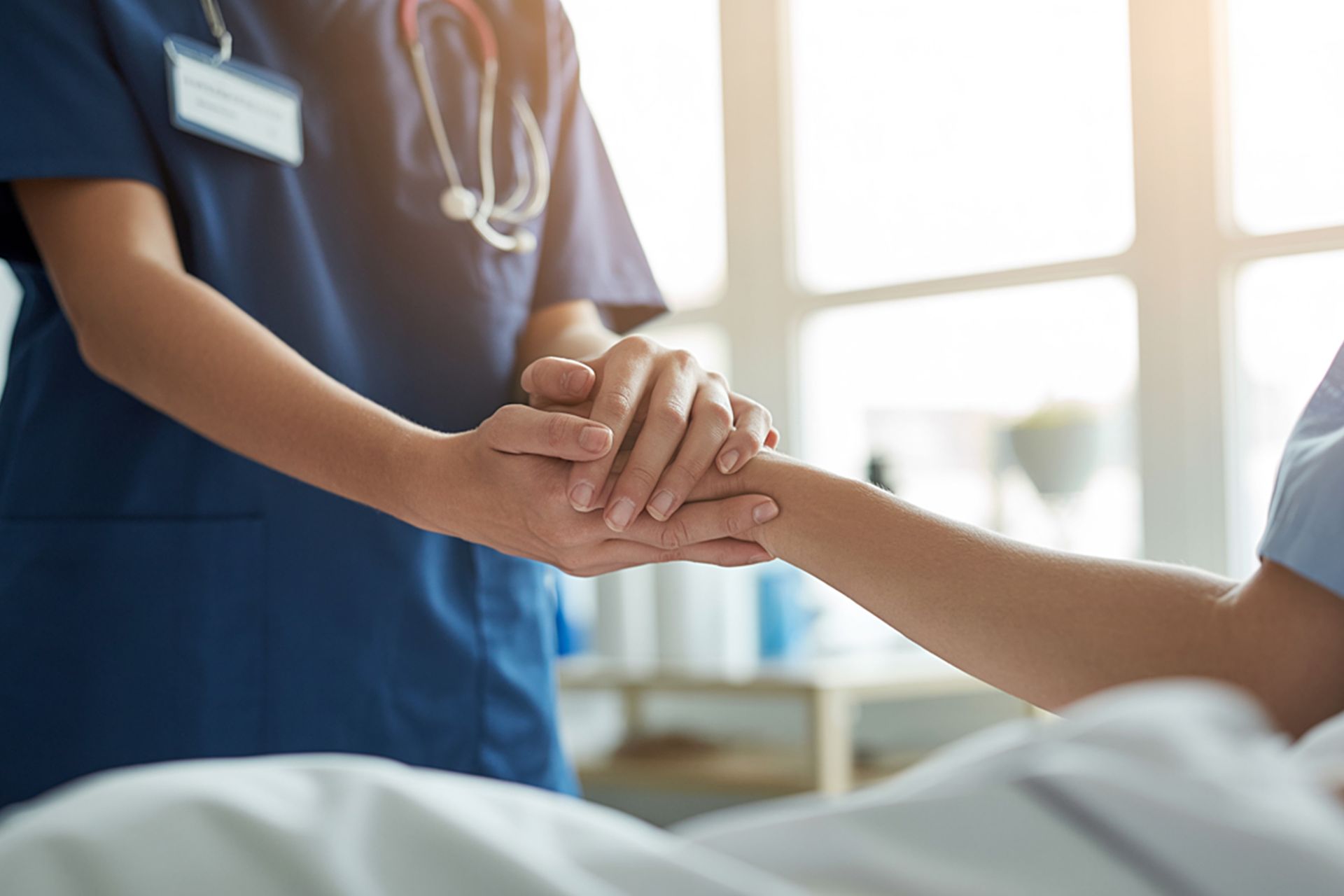 A healthcare worker in blue scrubs gently holding a patient's hand in a hospital setting.