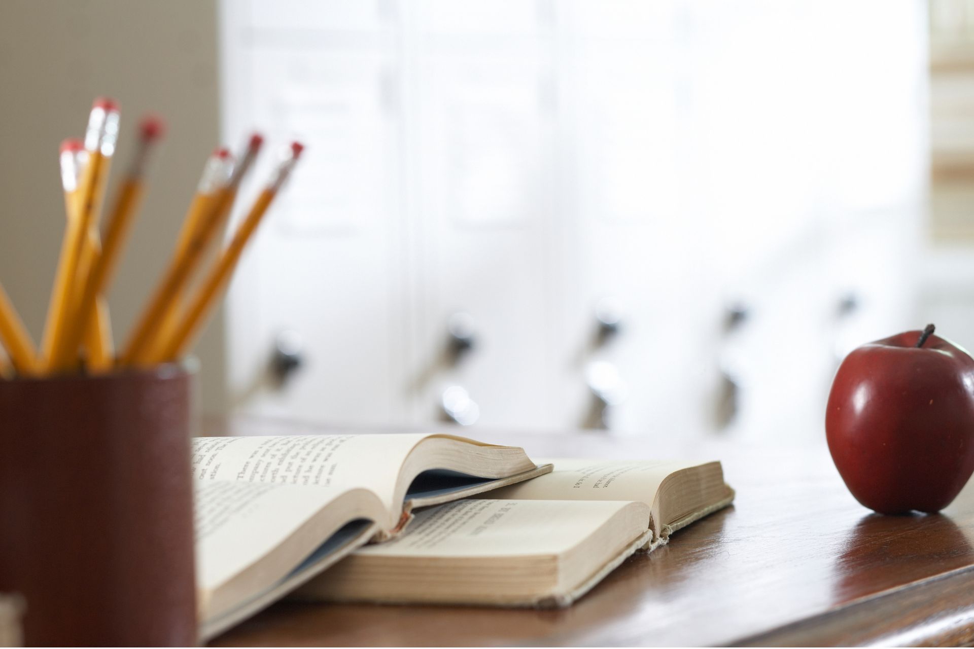An open book, a cup of sharpened pencils and a red apple sit on a school desk.