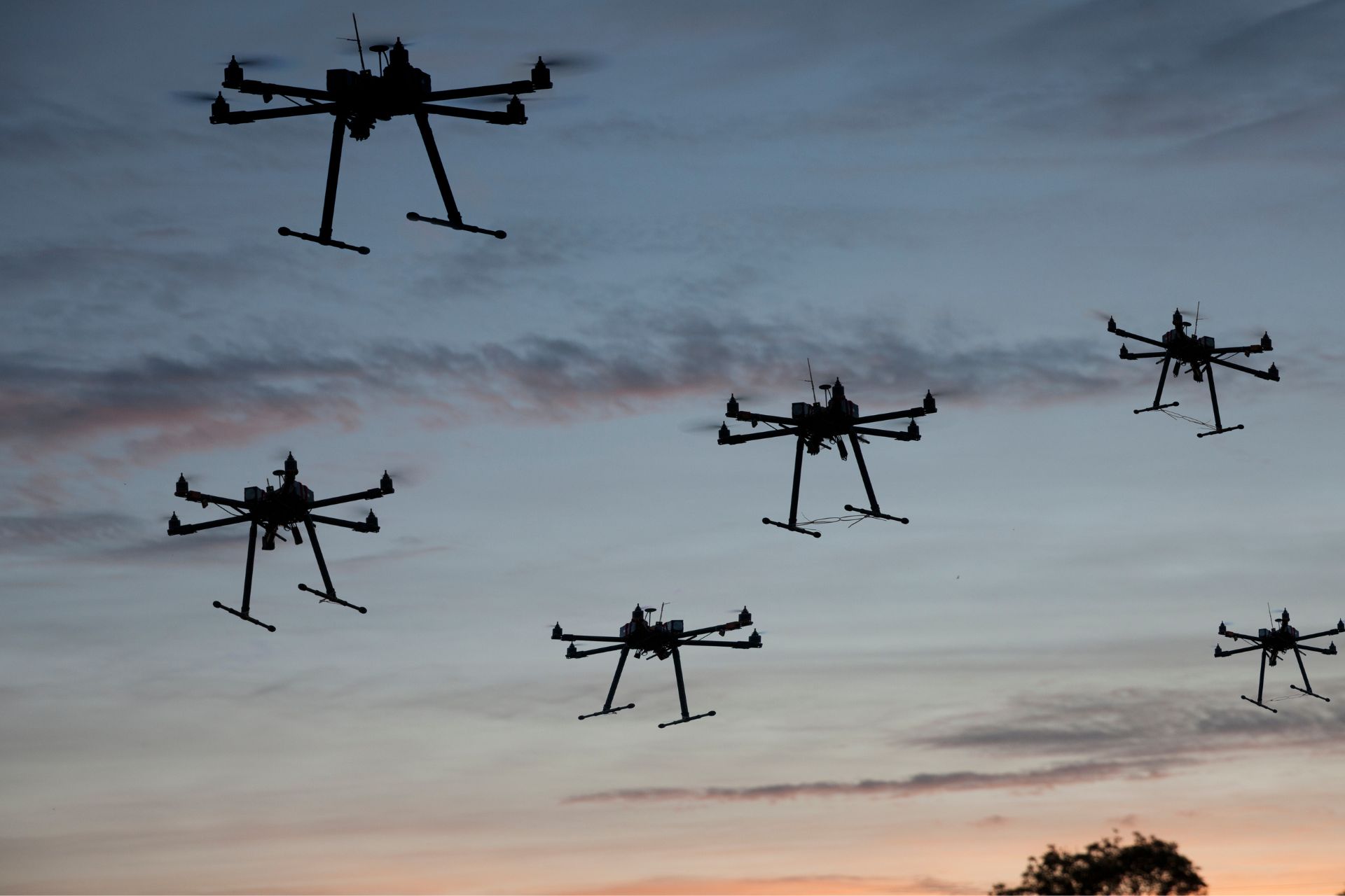 Several black multirotor drones hovering against a dusky evening sky