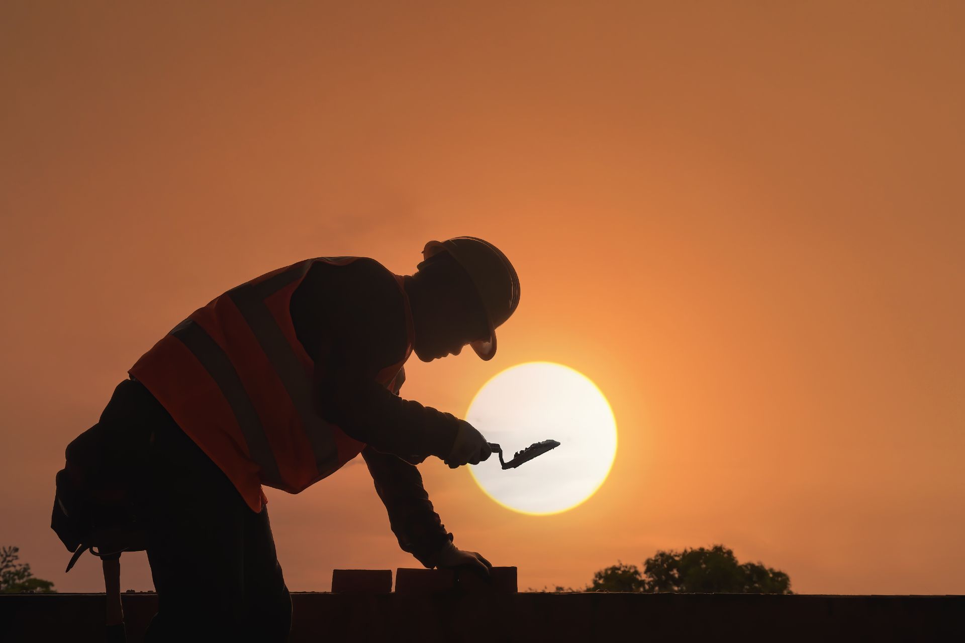 Silhouette of a construction worker crouched against a setting sun and orange sky