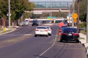 Cars drive down a two-lane street past a parked black sedan, with an orange “Utility Work” sign and traffic barrels along the right curb and an overpass in the background.