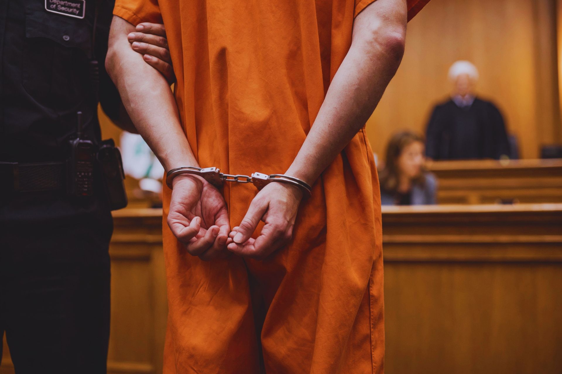 A defendant wearing an orange jumpsuit stands with their hands handcuffed behind their back in a courtroom.