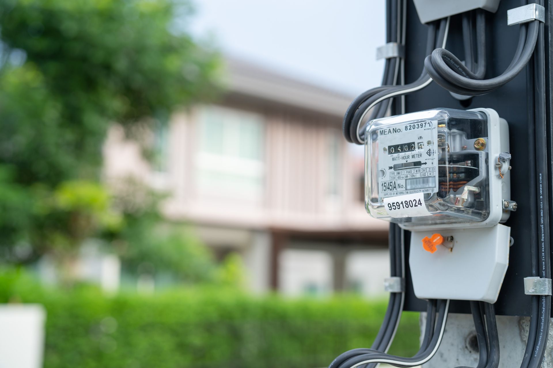 An electric meter mounted on a utility box outside a home, with wiring below it and a blurred house and green lawn in the background.
