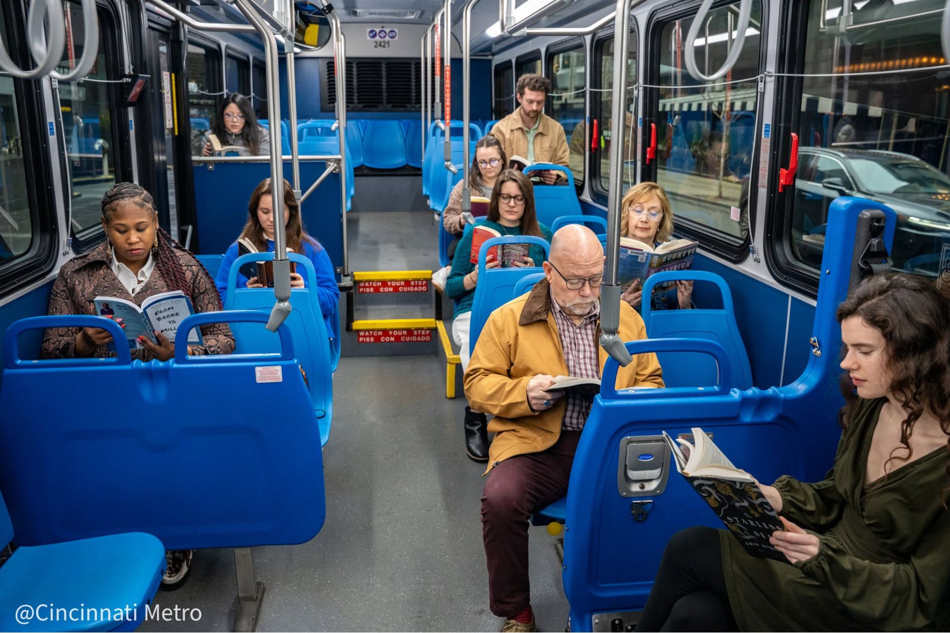 Passengers sit on blue seats inside a Cincinnati Metro bus, some reading or looking at their phones.