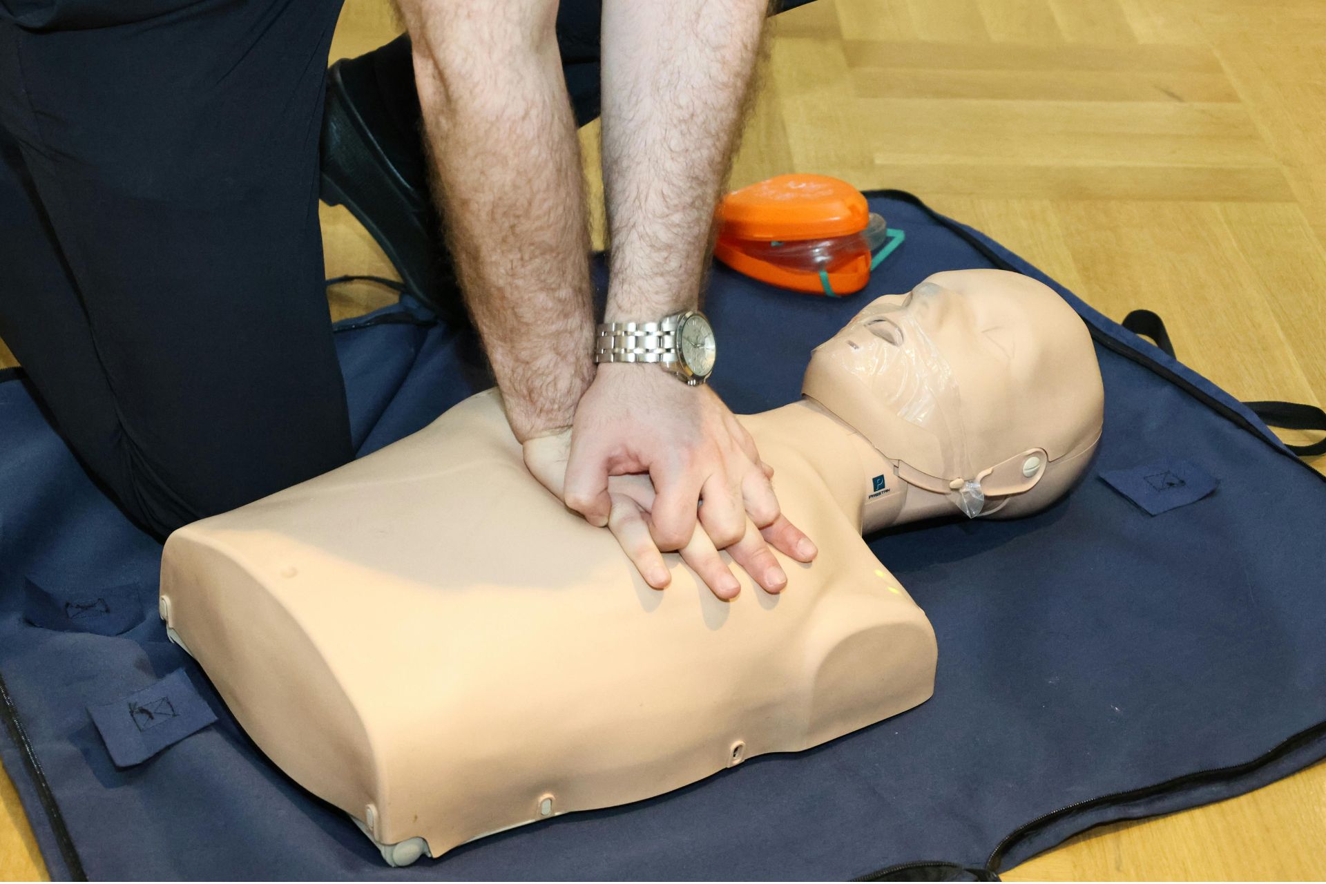 Hands perform chest compressions on a CPR training manikin lying on a blue mat during a training session.