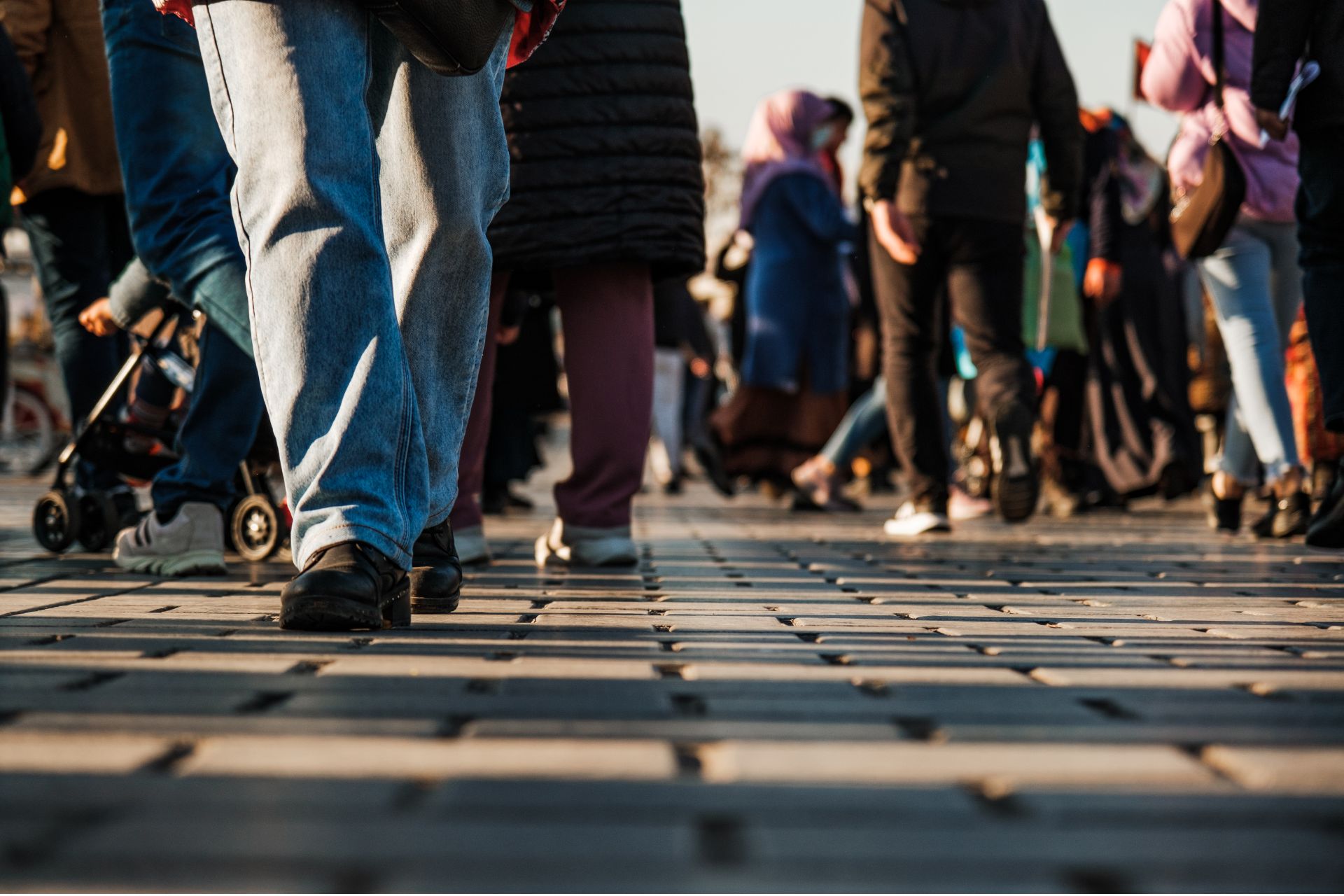 People walking along a sunlit city sidewalk, shown from knee level, including a stroller at left.