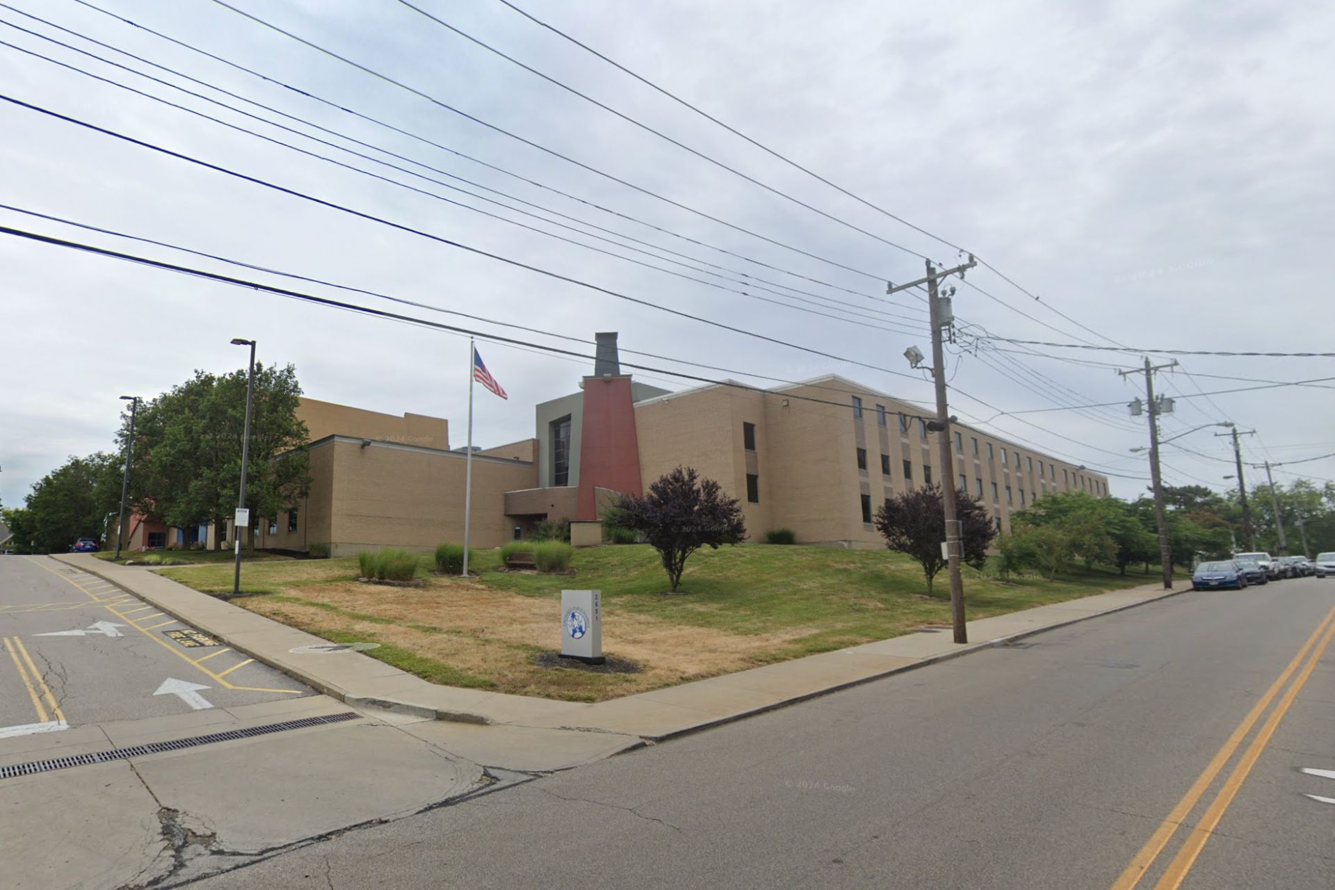 Brick school building with a grassy lawn, flagpole and utility poles along a street.
