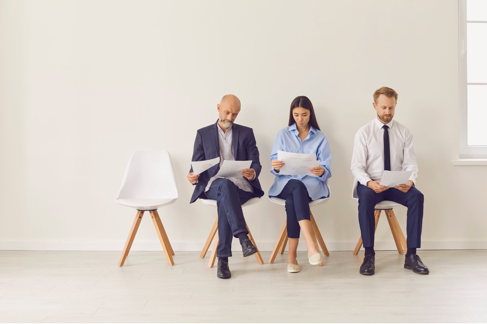 Three job seekers in business attire sitting in a waiting room holding folders, with an empty chair beside them.