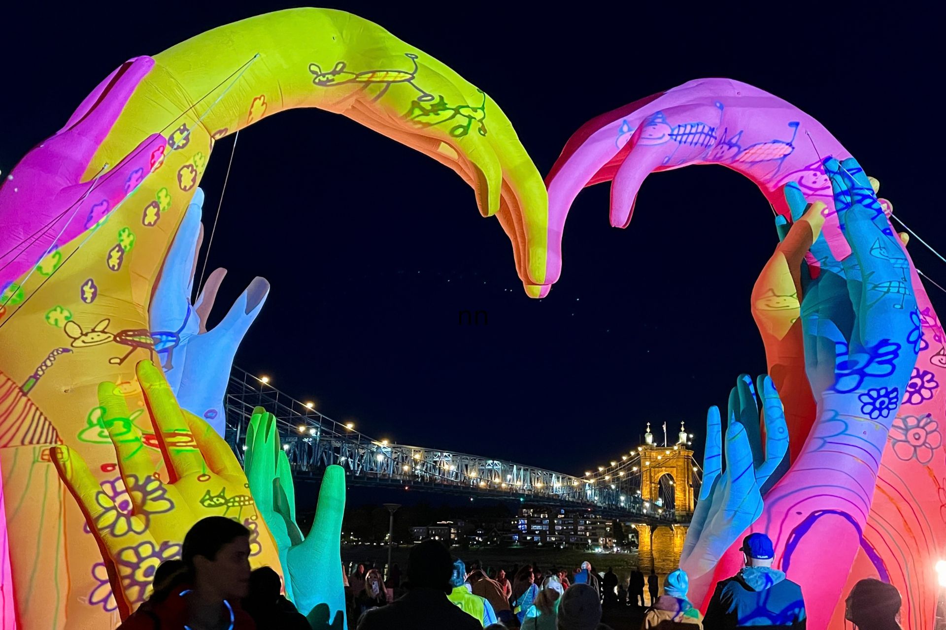 A brightly colored, illuminated sculpture of two giant hands forming a heart shape at night, with a crowd of people gathered below.