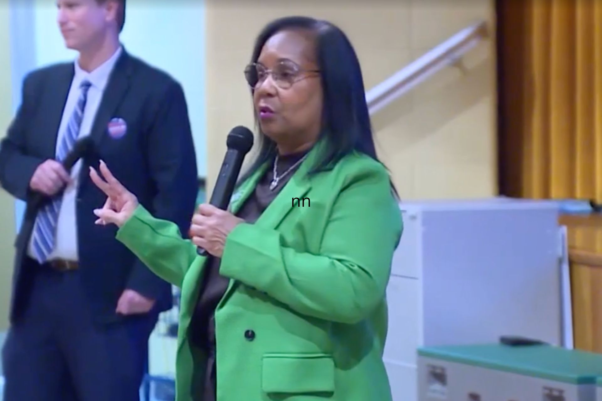 Woman in a green blazer speaks into a microphone and gestures while a man in a dark suit stands beside her