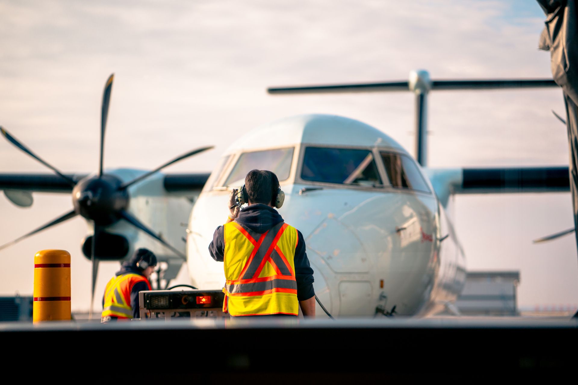 Airport ground crew worker wearing a high-visibility safety vest on the tarmac next to a twin-turboprop airplane.