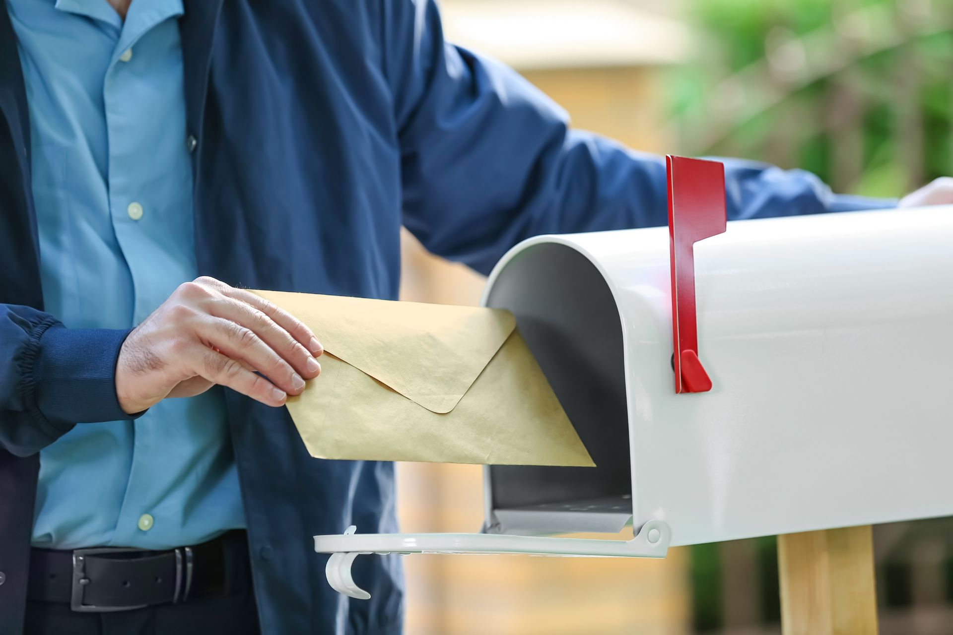 A person in a blue jacket slides a tan envelope into a white curbside mailbox with its red flag raised.