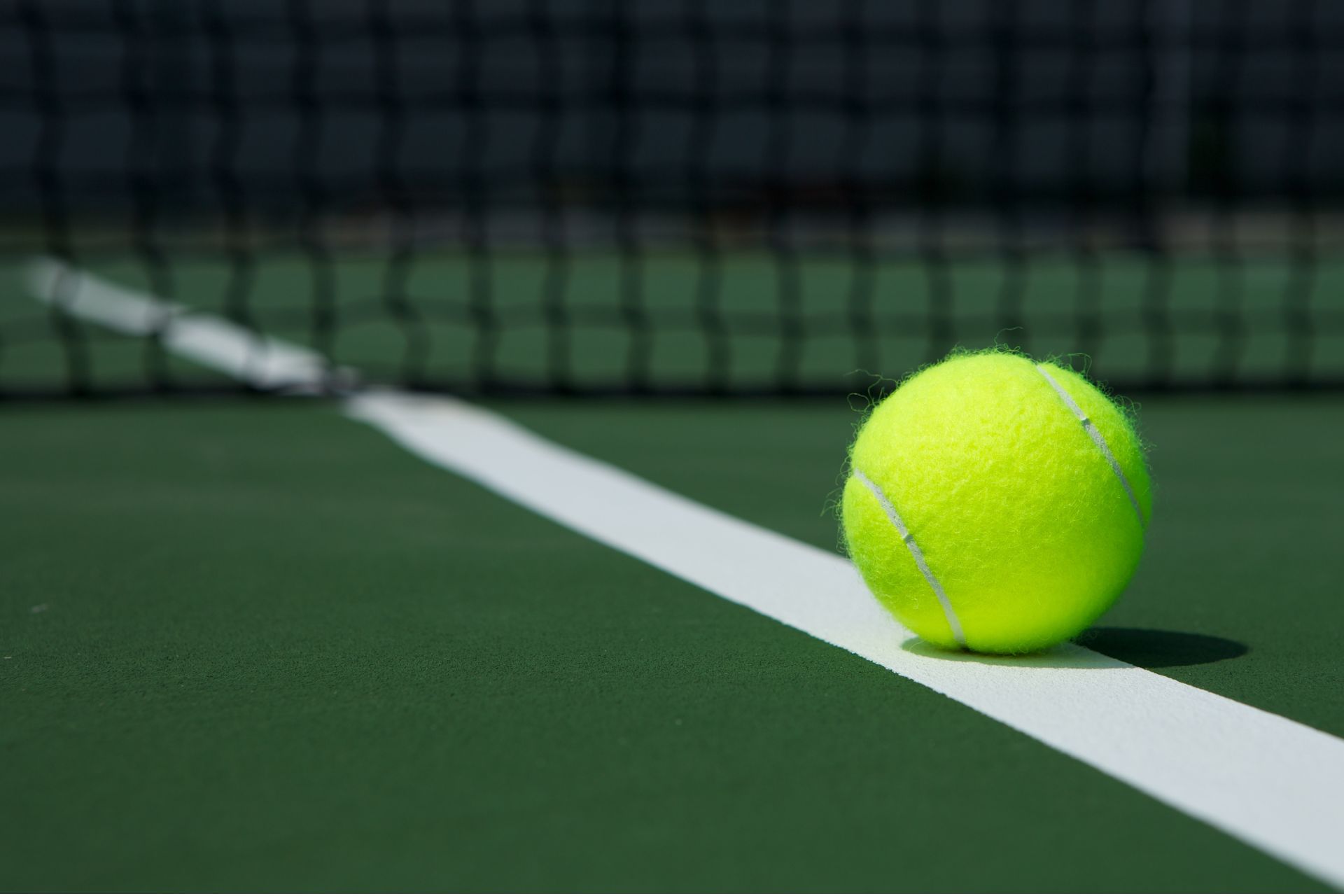 Yellow-green tennis ball resting on the white boundary line of a dark green tennis court.
