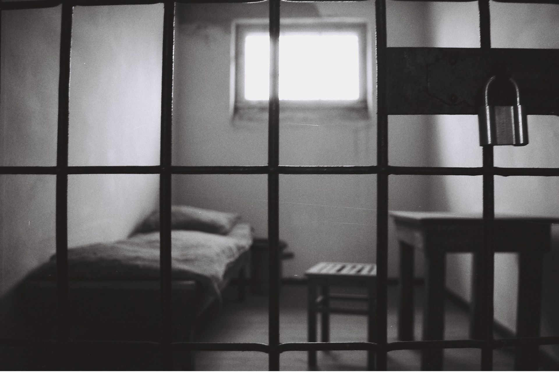 Black-and-white photo of a jail cell viewed through bars, with a bed, a small table and a padlock hanging on the bars.