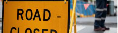 A bright yellow "ROAD CLOSED" barricade sign standing on a city street beside a road construction worker in high-visibility safety gear.