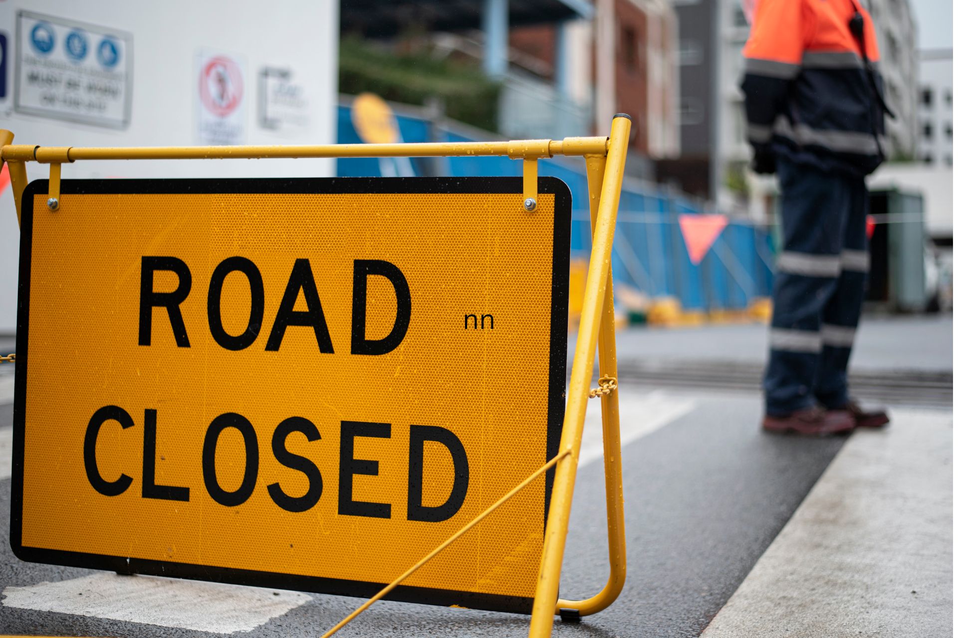 A bright yellow "ROAD CLOSED" barricade sign standing on a city street beside a road construction worker in high-visibility safety gear.