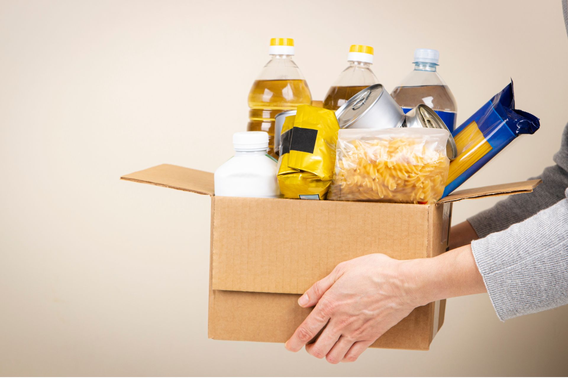 Hands holding a cardboard box filled with canned goods, pasta, and bottles of cooking oil