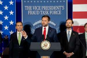 A man speaks at a podium in front of a backdrop reading “The Vice President’s Fraud Task Force,” with men in suits standing on either side and American flags behind them.