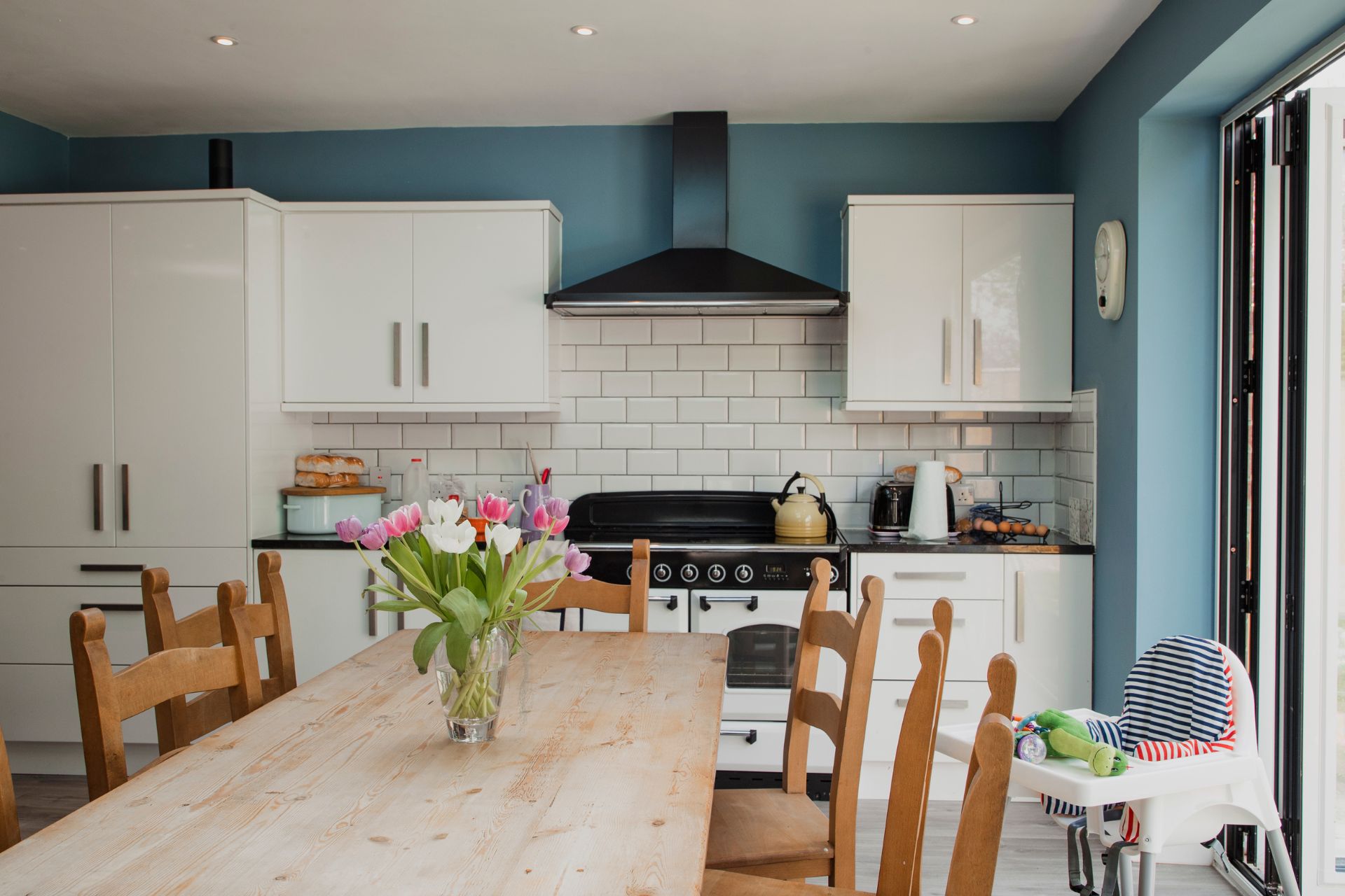 A bright kitchen and dining area with white cabinets, a wooden table and chairs, and a vase of flowers.