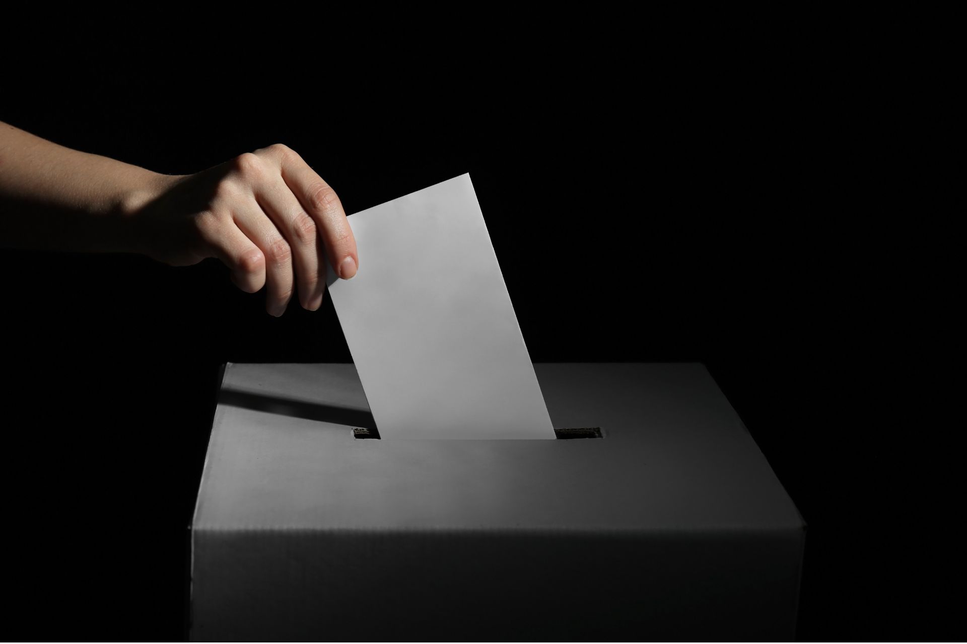 A hand drops a paper ballot into a ballot box against a dark background.