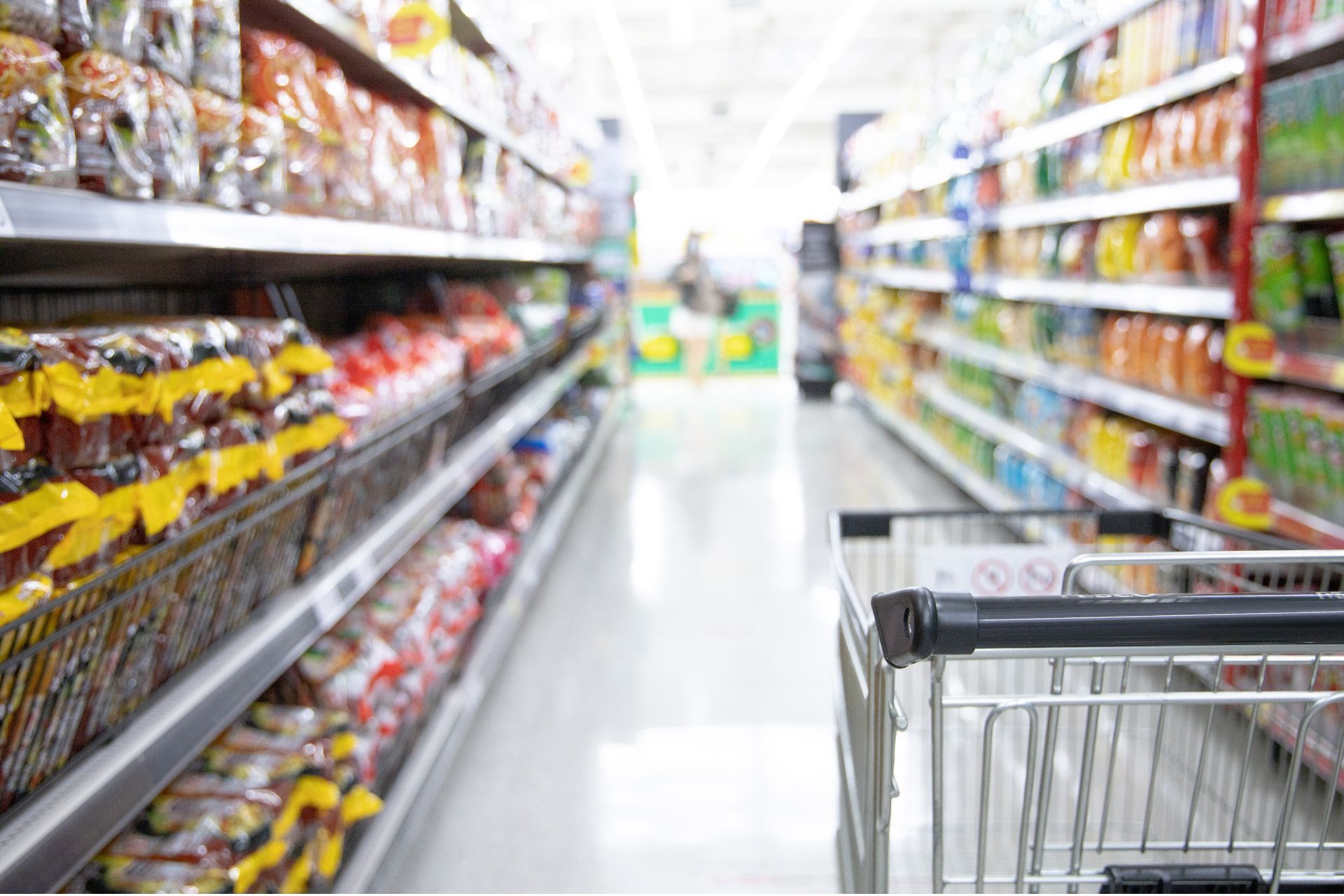 A grocery store aisle lined with shelves of packaged food, with a shopping cart in the foreground.