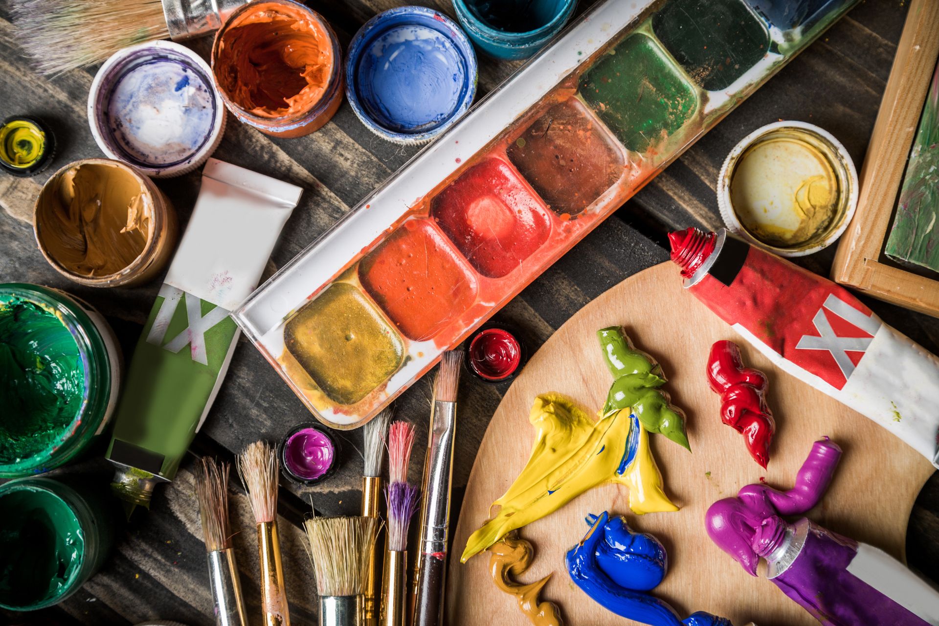 Overhead view of painting supplies, including a watercolor palette, tubes of paint and brushes, on a wooden table.